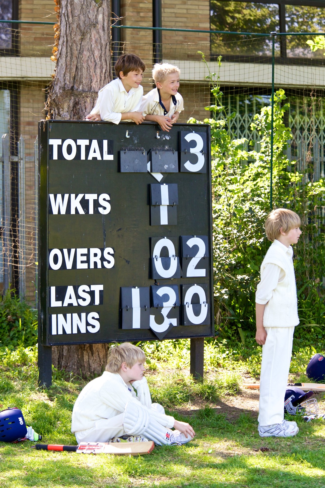 four boys watching a cricket match gathered around the scoreboard