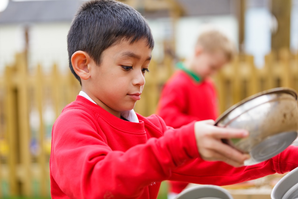 A young boy holds a metal bowl whilst playing outside.